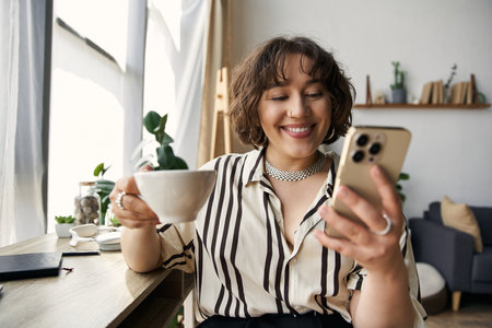 Bright light fills the chic apartment as a young woman smiles, sipping coffee and using her phone.の写真素材