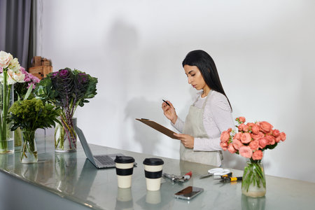 Brunette woman organizes flowers and takes notes in her vibrant flower shop, ready for customers.