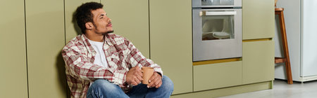 Handsome young man relaxes on the floor of a stylish kitchen while holding a warm drink.の写真素材