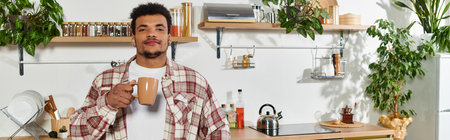 A young handsome man stands in a bright kitchen, holding a cup of coffee, surrounded by greenery.の写真素材
