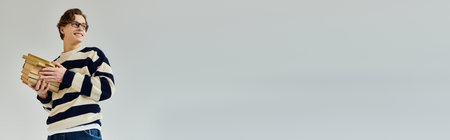 A young man in a striped sweater smiles while carrying books, showcasing college life in a studio, bannerの写真素材