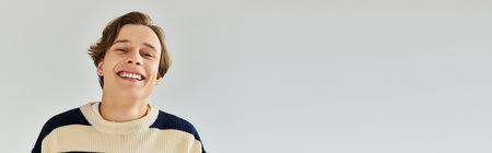 A cheerful young student wears a striped sweater while enjoying his college experience in a studio, bannerの写真素材