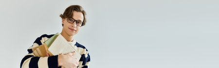 A young man confidently models a striped sweater while holding books in a studio, bannerの写真素材