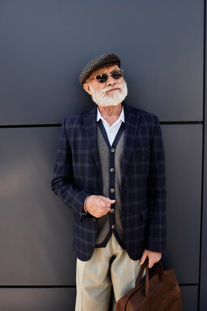 A dapper senior gentleman with a beard stands confidently by a contemporary structure, embracing autumn vibes.の写真素材