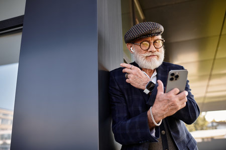 A fashionable senior man with a beard stands by a building, engaged in a phone conversation,...