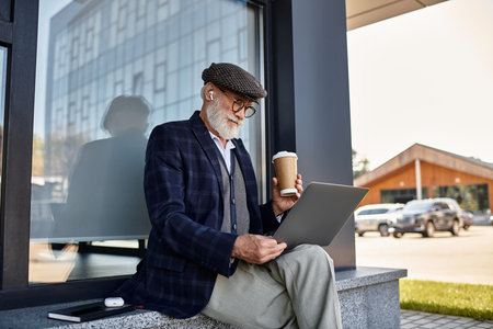 A fashionable senior man sits near a contemporary structure, sipping coffee and focused on his laptop in autumn.の写真素材