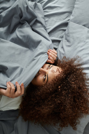 A woman peeks out from beneath gray blankets, enjoying a playful moment in bed.の写真素材
