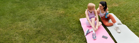 Two young women share smiles and laughter while relaxing on yoga mats in a green park.の写真素材
