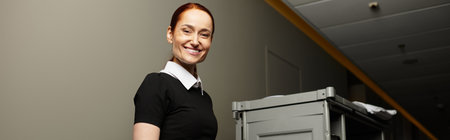 Beautiful young woman stands confidently in a bright office corridor with a cart.