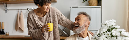 A woman handily smiles at her partner while holding a yellow mug in a warm kitchen setting.の写真素材