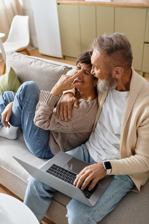 Mature couple shares laughter and affection while using a laptop on their comfortable sofa.の写真素材