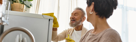 Cheerful senior couple shares laughter while cleaning their sunny kitchen on a joyful afternoon.の写真素材