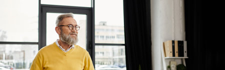 Handsome mature man with a beard stands in a stylish indoor space filled with light.の写真素材