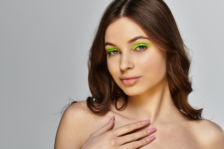 Young woman showcases vibrant green eyeshadow and flawless makeup against a grey backdrop.