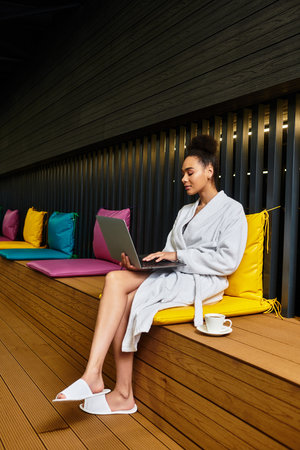 A young chic woman in a spa gown enjoys tea while working on her laptop.