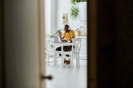 Brightly lit kitchen with a young african american man engaged in remote work, showcasing nomadism.の写真素材