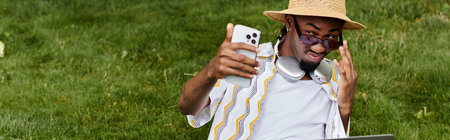 Handsome young man in a straw hat smiles while using a laptop and phone in a green park.の写真素材
