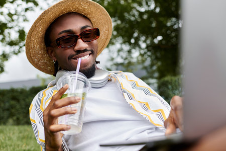 Relaxing in a lively park, a young man works on his laptop while sipping a refreshing drink.の写真素材