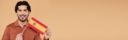 Brunette man smiles with a small Spanish flag on a warm, neutral background.の写真素材
