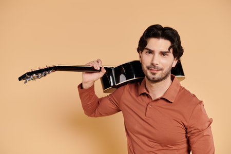 A young brunette man smiles gently while holding a guitar on his shoulder in a cozy setting.