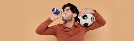 A young handsome man balances a soccer ball on his shoulder while drinking a refreshing beverage.の写真素材