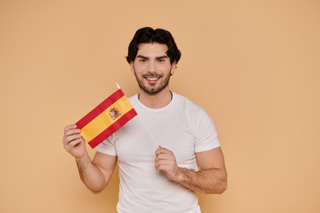 Handsome young man smiles while displaying a small Spanish flag, radiating joy and pride.の写真素材