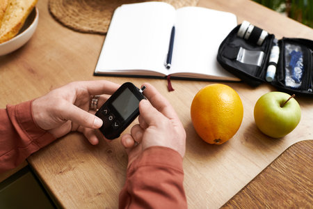 A young man checks his blood sugar levels at home surrounded by healthy snacks and planning tools.の写真素材