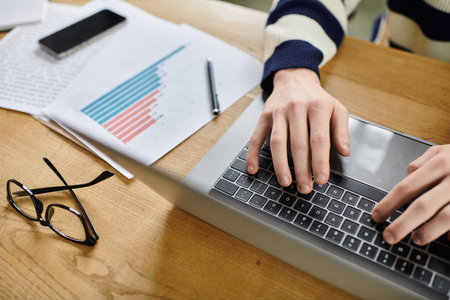 A person types on a laptop while surrounded by office materials and reports.