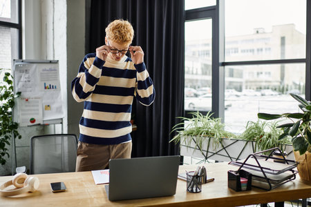 Young redhead man in striped sweater actively engages with his laptop in a sleek office setting.