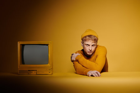 A young man with red hair presents a fashionable winter look near vintage television against a...