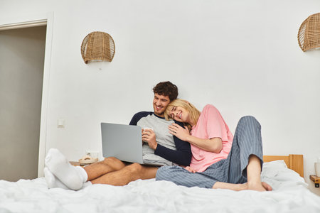 Two men relax on a bed, sharing laughter and warmth while browsing their laptop in a cozy room.の写真素材