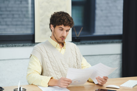 Man with curly hair in a sweater vest reviews documents at a modern office desk.
