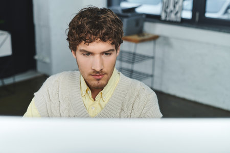 Curly-haired man in a sweater vest engages with his computer in a fresh corporate workspace.