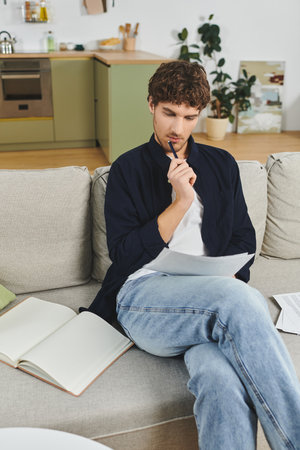 Young man sits thoughtfully on the couch in a chic apartment while reviewing notes.