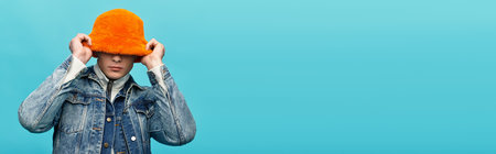 A handsome young man adjusts his striking orange hat while standing before a vivid blue backdrop.の写真素材