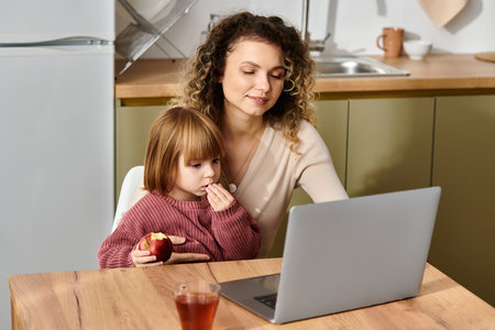 Curly-haired mother and her daughter engage with a laptop while enjoying a snack at home.の写真素材