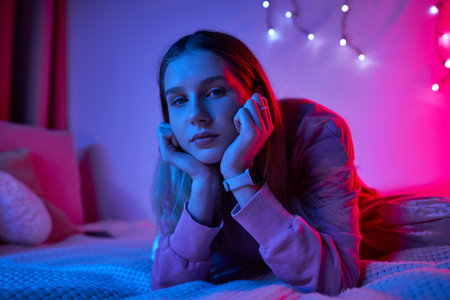 A teenage girl relaxes in her stylish room, surrounded by colorful lights in the evening.