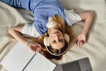 Teen girl lies on her bed in a cozy room, enjoying music while studying with a laptop.の写真素材