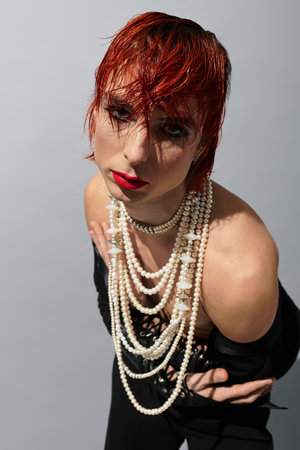 A unique young man flaunts bold red hair and layered pearl necklaces against a plain background.