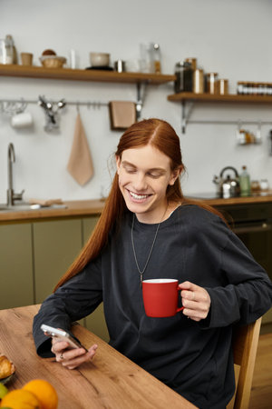 Charming young man with long hair sips coffee in his stylish apartment, checking his phone.の写真素材