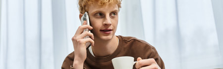 A handsome young man with red hair talks on his phone while sipping coffee at home.