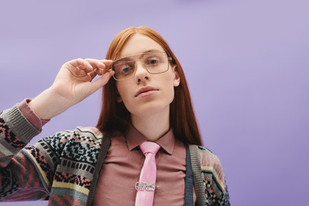 Young queer man with long red hair dons glasses and a tie, showcasing modern fashion flair.のeditorial素材