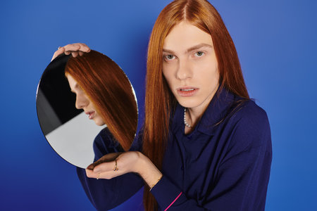 Handsome young man with long red hair holds a mirror, showcasing his bold, stylish essence.のeditorial素材