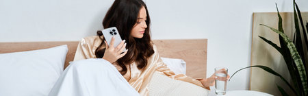 Young woman in a silk robe relaxes in bed with her smartphone and a glass of water.のeditorial素材