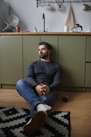 A handsome man sits peacefully on the kitchen floor, lost in thought while sipping a drink.の写真素材