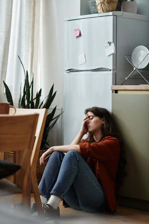 In a cozy kitchen, a woman leans against the wall, her face showing inner turmoil and sadness.の写真素材