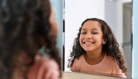 A young girl with curly hair smiles joyfully while looking in the mirror at a dental clinic.の素材