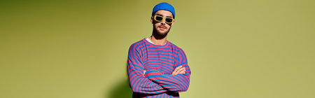 A confident young man poses in a trendy outfit, wearing vibrant sunglasses against a green backdrop,...
