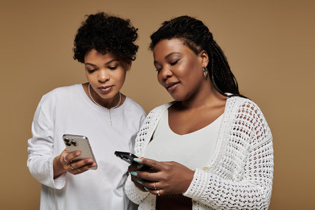 Two stylish women share a cozy moment, smiling and using their phones.