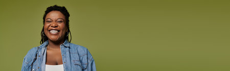 Stylish african american woman in vibrant denim attire with long hair posing on olive backdrop.